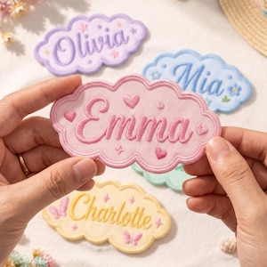 May include: A pink embroidered cloud-shaped patch featuring the name Emma in cursive script, accented with small hearts and star motifs. The patch has a scalloped border and is held against a background of other colorful name patches.
