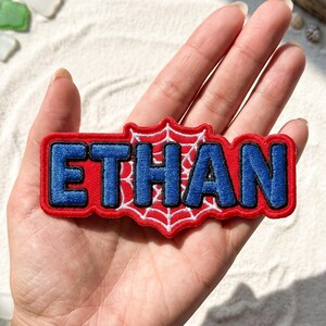 May include: A rectangular embroidered patch featuring the name ETHAN in bold blue letters with a black outline. The background of the patch is a red and white spiderweb design, and the entire piece is bordered by a thick red stitched edge.