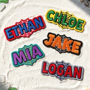 May include: Five embroidered name patches featuring spiderweb designs. The patches display the names ETHAN in blue, CHLOE in green, JAKE in orange, MIA in purple, and LOGAN in red. Each name is outlined in a contrasting color with a web background.