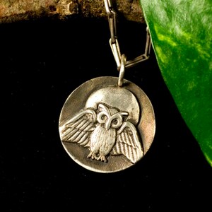 May include: Silver owl pendant necklace with a detailed owl design on a round disc. The owl is depicted with its wings spread, and the disc has a textured surface.