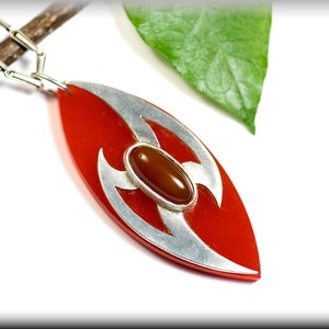 Könnte beinhalten: Eine rote Anhängerkette mit silberfarbenem Design und einem braunen ovalen Stein in der Mitte. Der Anhänger befindet sich an einer silbernen Kette. Ein grünes Blatt und ein Zweig sind im Hintergrund.