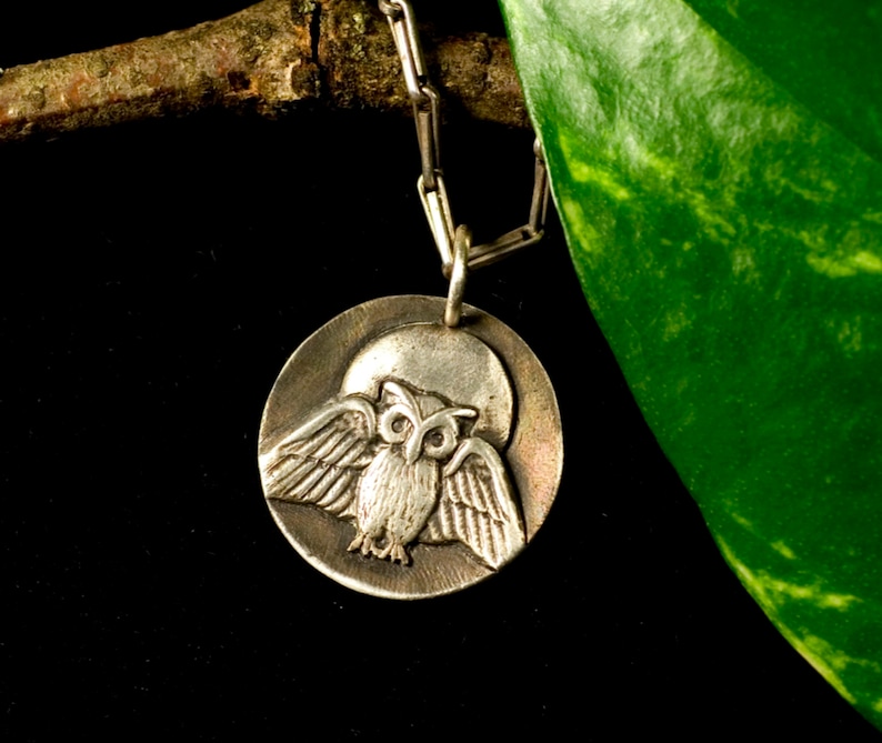 May include: Silver owl pendant necklace with a detailed owl design on a round disc. The owl is depicted with its wings spread, and the disc has a textured surface.