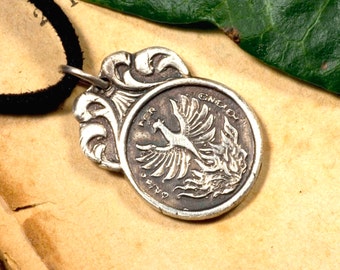 May include: A silver pendant necklace with a detailed design featuring a phoenix rising from flames. The pendant is on a black cord and is resting on a piece of aged paper.