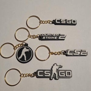 Puede incluir: Cuatro llaveros con anillas y cadenas de metal dorado. Uno presenta el texto CSGO, otro dice COUNTER STRIKE 2, otro muestra CS2 y otro exhibe la silueta de una persona sosteniendo un rifle junto al texto CSGO. Todos son en blanco y negro.