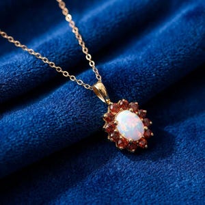May include: A gold chain necklace featuring an oval-shaped pendant. The pendant has a central white iridescent oval stone surrounded by a halo of small, deep red faceted stones arranged in a floral-like pattern set in a gold metal frame.