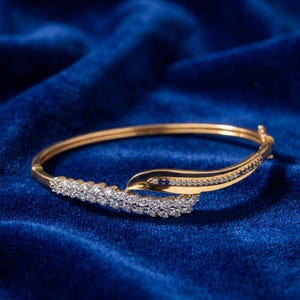 Op de afbeelding: Een gouden scharnierende armband met een gebogen ontwerp, versierd met talloze kleine, heldere, rondgeslepen stenen. De armband heeft een gepolijste gouden afwerking en een veilig sluitingsmechanisme, met een doorlopende band van fonkelende stenen.