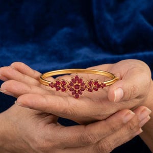 May include: A gold bangle bracelet featuring a central floral-inspired motif adorned with multiple marquise-cut and square-cut red rubies. The metal band is smooth and circular, with the gemstone arrangement centered on the top portion of the piece.