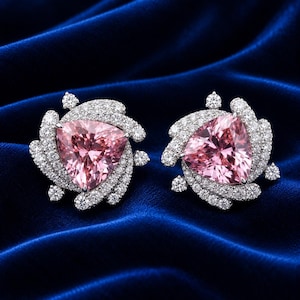 Puede incluir: Un par de pendientes de botón con una gran piedra central rosa de corte triangular, rodeada por un halo en espiral de pequeñas piedras claras engastadas en pavé. El engaste de metal plateado crea un contraste brillante con las gemas rosas.