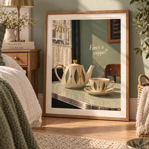 Op de afbeelding: Een rechthoekige ingelijste kunstdruk met een theepot en theekop in mid-century stijl op een tafel. De afbeelding toont de tekst "Fancy a cuppa?" in een cursief lettertype. De theepot en kop hebben een crèmekleurige basis met blauwgroene en gouden bladpatronen.