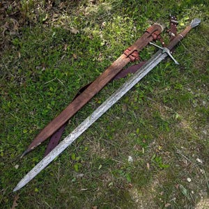 Può includere: Una spada lunga con lama in acciaio Damasco a motivi, guardia in metallo a croce e impugnatura in legno marrone con pomolo arrotondato. Accanto alla spada si trova un fodero in pelle marrone abbinato con intricati disegni floreali.
