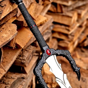 Puede incluir: Una espada decorativa con mango negro texturizado, pomo en forma de calavera y una piedra roja. La guarda tiene forma de garras negras que sostienen una piedra roja, y la hoja plateada muestra un texto grabado en una caligrafía.