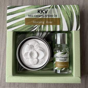 Op de afbeelding: Een KKV-cadeauset met een witte keramische bloemendiffuser in een rond metalen blik en een flesje van 10ml Morning Rose olie. De verpakking heeft een groen palmbladpatroon en de tekst KKV Mini Ceramic & 10ml Oil Morning Rose.