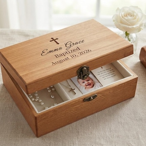 May include: A rectangular wooden keepsake box with a hinged lid and a metal latch. The lid is engraved with a cross, the name Emma Grace, and the text Baptized August 10, 2026. Inside, the box contains a silver cross necklace and a baby photo.