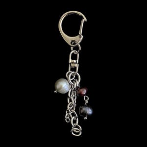 Può includere: Un portachiavi in metallo color argento con un anello circolare e un moschettone. Alla catena sono appese tre perle distinte: una grande perla bianca, una piccola perla marrone rossastro scuro e una perla grigio scuro di medie dimensioni.