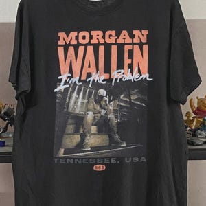 Peut inclure: Un t-shirt noir à manches courtes avec le texte orange MORGAN WALLEN, le texte blanc en écriture manuscrite I'm the Problem et une impression photographique d'une personne assise sur des marches. Sous l'image, le texte TENNESSEE, USA et 865.