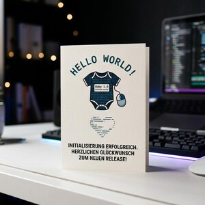 Könnte beinhalten: Eine weiße Grußkarte mit dem Text HELLO WORLD! in Dunkelblau, einer Grafik eines dunkelblauen Babybodys mit der Aufschrift Baby 1.0, der mit einer Computermaus verbunden ist, einem Herz aus Code und dem Text INITIALISIERUNG ERFOLGREICH.