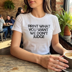 May include: A white short-sleeved t-shirt featuring the black text PRINT WHAT YOU WANT WE DON'T JUDGE centred on the chest. The garment has a crew neckline and a standard fit, constructed from a plain white fabric with bold black lettering.