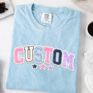 May include: A light blue t-shirt featuring the word CUSTOM in varsity-style letters with a chenille texture. The letters are colored in pink, cream, blue, and purple. Below the text are three small stars in navy, pink, and light purple.