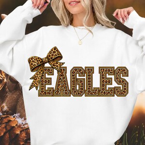 May include: A white long-sleeve sweatshirt featuring the word EAGLES in large, block-style letters with a brown and black leopard print pattern. A matching leopard print bow is positioned to the left of the text, adding a decorative element.