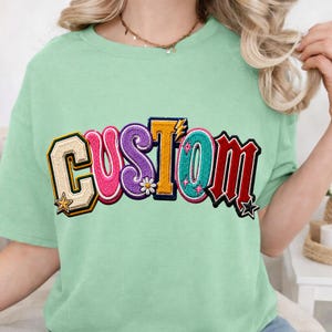 May include: A light green short-sleeve t-shirt featuring the word CUSTOM in large, multi-coloured, textured varsity-style letters. The letters are decorated with small star, flower, and lightning bolt icons in shades of pink, purple, yellow, and teal.