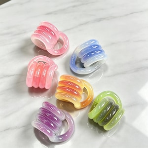 May include: A collection of five translucent, coiled hair clips in various colors including pink, blue, orange, purple, and green. Each clip features a rounded, multi-loop design with a smooth, glossy finish, arranged in a cluster on a surface.