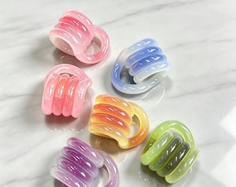 May include: A collection of five translucent, coiled hair clips in various colors including pink, blue, orange, purple, and green. Each clip features a rounded, multi-loop design with a smooth, glossy finish, arranged in a cluster on a surface.