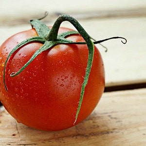 May include: A single, ripe red tomato with a green stem and water droplets on the surface. The tomato is sitting on a wooden surface.