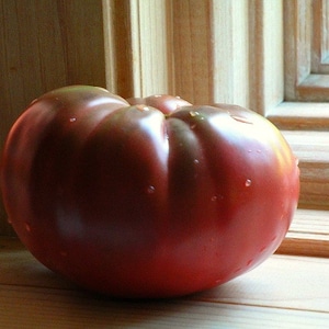 May include: A large, deep red heirloom tomato with a slightly bumpy surface. The tomato is sitting on a wooden surface.