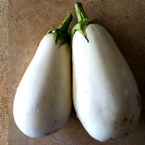 May include: Two white eggplants with green stems. The eggplants are sitting on a brown surface.