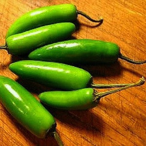 May include: Five green jalapeno peppers on a wooden surface. The peppers are all different sizes and shapes, and some have stems.