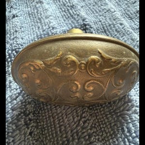 May include: An oval-shaped brass door knob featuring a raised, intricate scrollwork pattern across its surface. The metal has a muted, aged finish with darker tones settled into the recessed areas of the embossed decorative design.