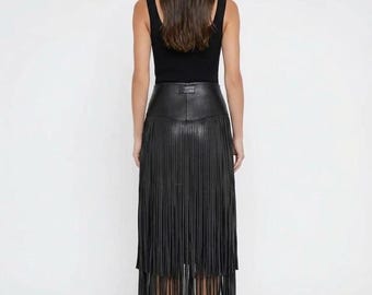 Puede incluir: Una camiseta negra sin mangas combinada con una falda midi negra que presenta un canesú de cuero de cintura alta y un dobladillo largo de flecos en capas que se extiende por debajo de la rodilla. La falda tiene una sección superior de cuero liso.