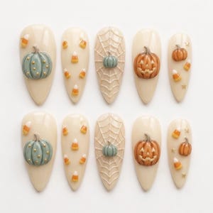 May include: A set of ten almond-shaped press-on nails with a cream base. Designs include sage green pumpkins with gold dots, orange candy corn, white spider webs with a small green pumpkin, and orange jack-o'-lanterns with gold star accents.