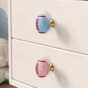 以下が含まれることがあります： 2段の引き出しが付いた白いナイトテーブルで、アメリカンフットボールの形をした装飾的な引き出しノブが2つ付いています。上のノブは水色に赤いレース、下のノブは薄いピンク色に赤いレースが施されており、どちらもゴールドトーンの金具で取り付けられています。