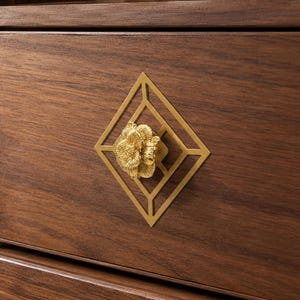May include: A dark brown wooden drawer featuring a gold-colored metal geometric diamond-shaped inlay centered around a three-dimensional gold floral knob. The drawer is part of a larger wooden cabinet structure with vertical fluted side detailing.