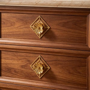May include: A dark brown wooden cabinet featuring two drawers with gold-toned metal hardware. Each drawer pull consists of a diamond-shaped frame surrounding a central, three-dimensional flower-shaped knob with detailed petals and a textured center.