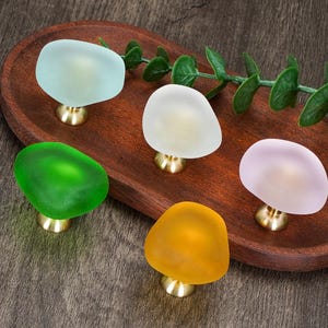 May include: Five cabinet knobs with smooth, rounded, frosted glass tops in light blue, white, bright green, soft pink, and yellow. Each knob features a small, circular, metallic gold-toned base that connects the glass top to the mounting surface.