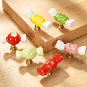 May include: A collection of six ceramic cabinet knobs shaped like wrapped candies. The knobs feature various colors including yellow, green, red, and pink, with some displaying white polka dot patterns or floral details, all mounted on brass-toned bases.
