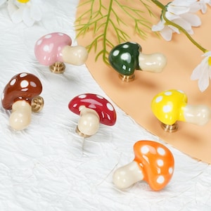 Puede incluir: Un juego de seis pomos para armarios de cerámica con forma de seta en varios colores, incluyendo rosa, verde oscuro, marrón, rojo, amarillo y naranja. Cada pomo tiene un sombrero brillante con lunares blancos y un tallo crema con base de latón.