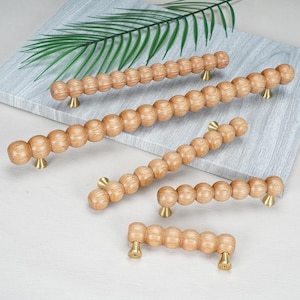 May include: A collection of five wooden cabinet pulls in varying lengths, each featuring a series of connected spheres with a natural light brown wood grain finish. Each pull is secured by two small, conical brass-toned metal mounting posts.