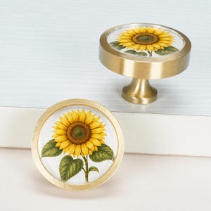 May include: Two round cabinet knobs featuring a brushed brass finish and a circular glass inlay. Each knob displays a detailed illustration of a vibrant yellow sunflower with green leaves centred on a white background.