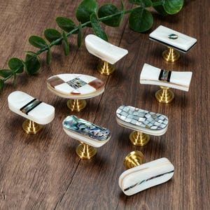 May include: A collection of eight cabinet knobs featuring white shell, iridescent abalone, and brown inlay patterns set on brass-toned metal bases. Each knob has a distinct geometric or elongated shape, showcasing natural textures and polished finishes.
