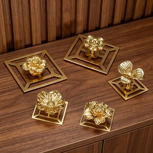 May include: Five gold-colored metal cabinet knobs, each featuring a three-dimensional flower design mounted on a square, open-frame base. The floral shapes vary in petal structure and size, showcasing a polished, reflective metallic finish.