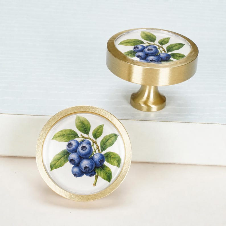 May include: Two round cabinet knobs with a brushed gold metal finish. The top surface of each knob features a detailed illustration of a cluster of blue blueberries with green leaves on a white background, protected by a clear glass cover.