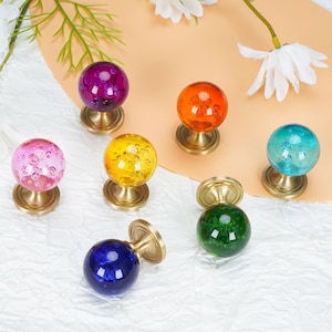May include: A collection of seven spherical cabinet knobs featuring translucent colored glass with internal bubble patterns. Each knob is attached to a circular brushed brass base. The colors include purple, orange, pink, yellow, teal, blue, and green.