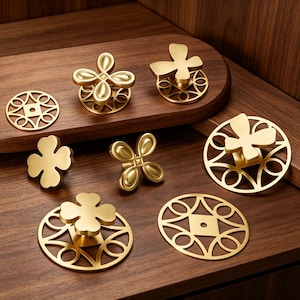 Op de afbeelding: Een collectie goudkleurige metalen kastknoppen en ronde achterplaten met ingewikkelde uitgesneden patronen. De set bevat klaverblad-vormen, vlinder-geïnspireerde ontwerpen en sierlijke geometrische filigraanschijven met een gladde afwerking.