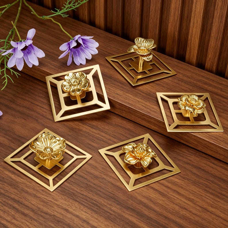 May include: Five gold-colored metal cabinet knobs, each featuring a square open-frame backplate with a central three-dimensional flower-shaped handle. The hardware has a polished metallic finish and a geometric design with a floral accent.