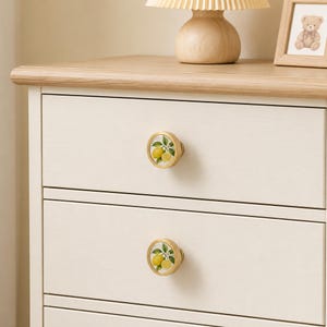 May include: Two round drawer knobs with a gold-toned metal rim and a white centre featuring a botanical illustration of yellow lemons with green leaves and stems, installed on a light-coloured wooden dresser with multiple drawers.