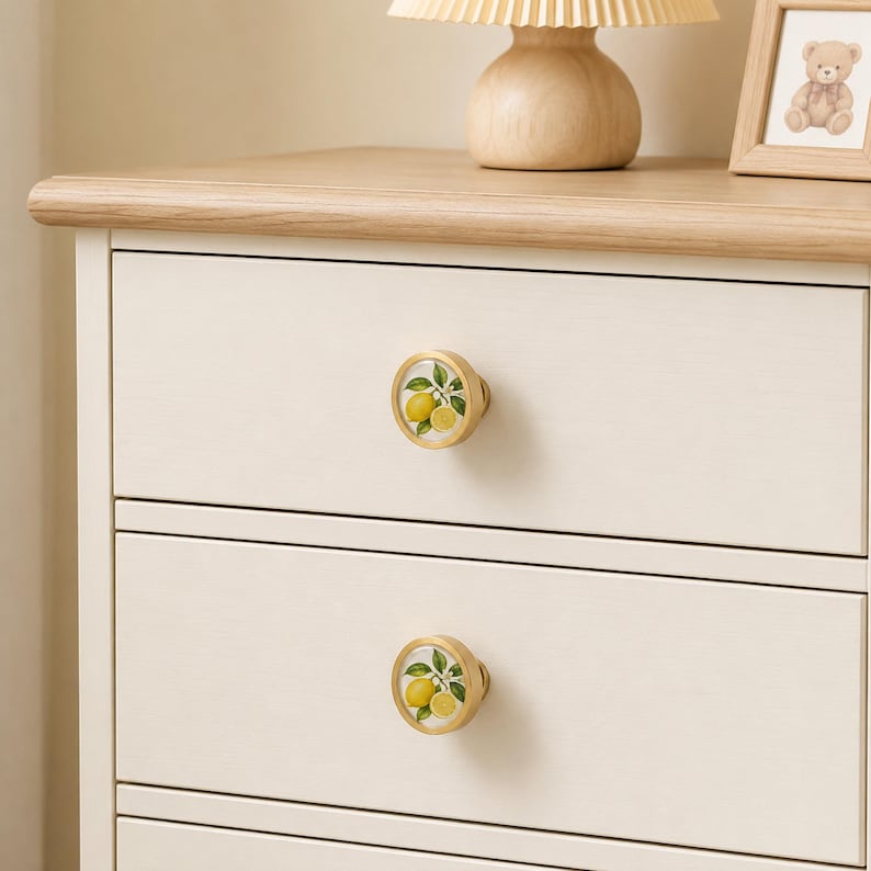 May include: Two round drawer knobs with a gold-toned metal rim and a white centre featuring a botanical illustration of yellow lemons with green leaves and stems, installed on a light-coloured wooden dresser with multiple drawers.