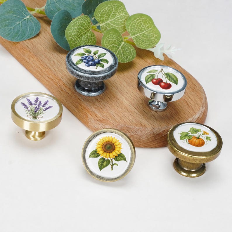 May include: Five round cabinet knobs with botanical illustrations on white ceramic inserts. The designs feature blueberries, cherries, lavender, a sunflower, and a pumpkin. The knobs have metallic finishes in silver, gold, and bronze tones.